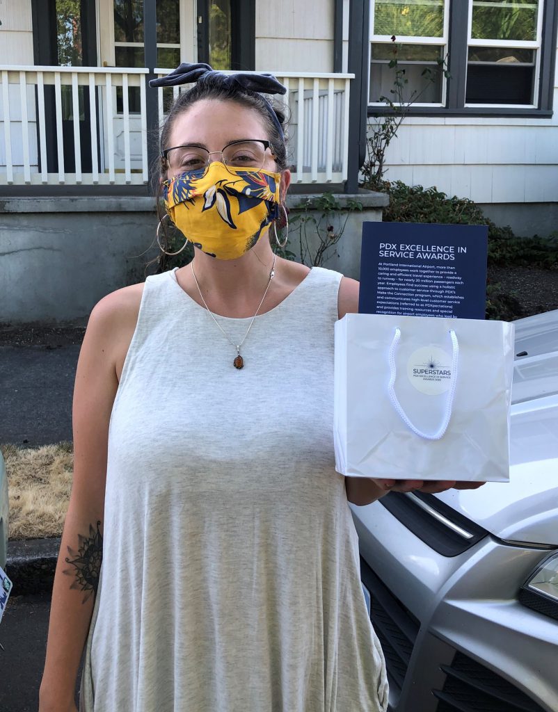 Autumn Daves, Blue Star Donuts employee, holding her PDX Excellence in Service award. She is standing in front of her house, wearing a yellow face covering and smiling with her eyes.