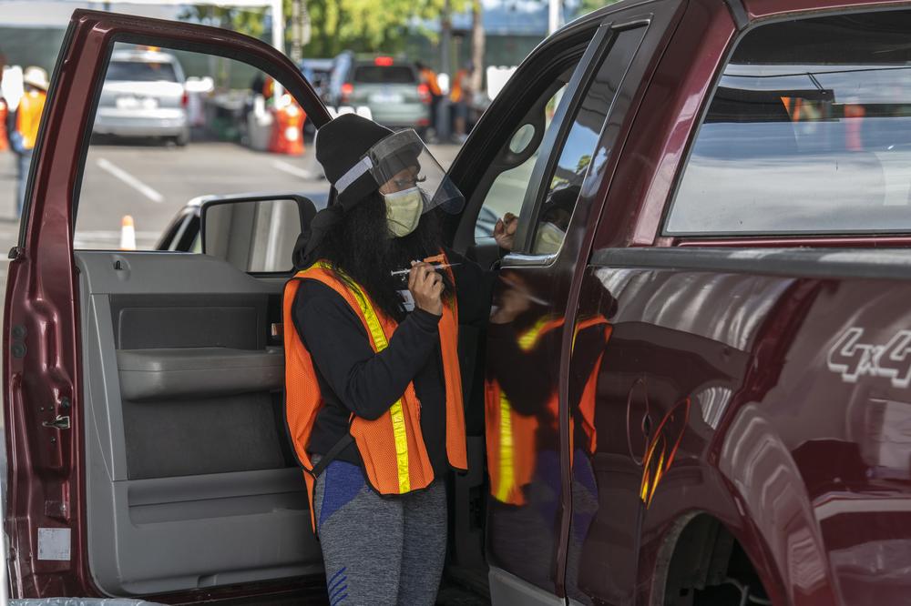 A woman in an orange safety vest and a mask and face shield holds a vaccine, while a drive in a red truck has their door open and is ready to get vaccinated