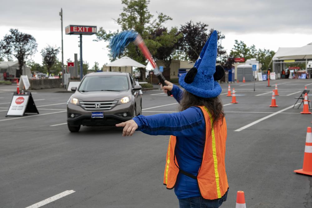 A woman wearing a blue shirt and orange safety vest wave a car forward into the PDX Red Lot
