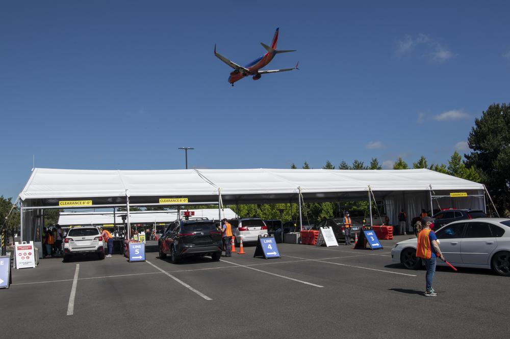 A jet plane flies overhead, while underneath a large white tent with six rows has vehicles underneath, where people are lining up to get vaccinated.