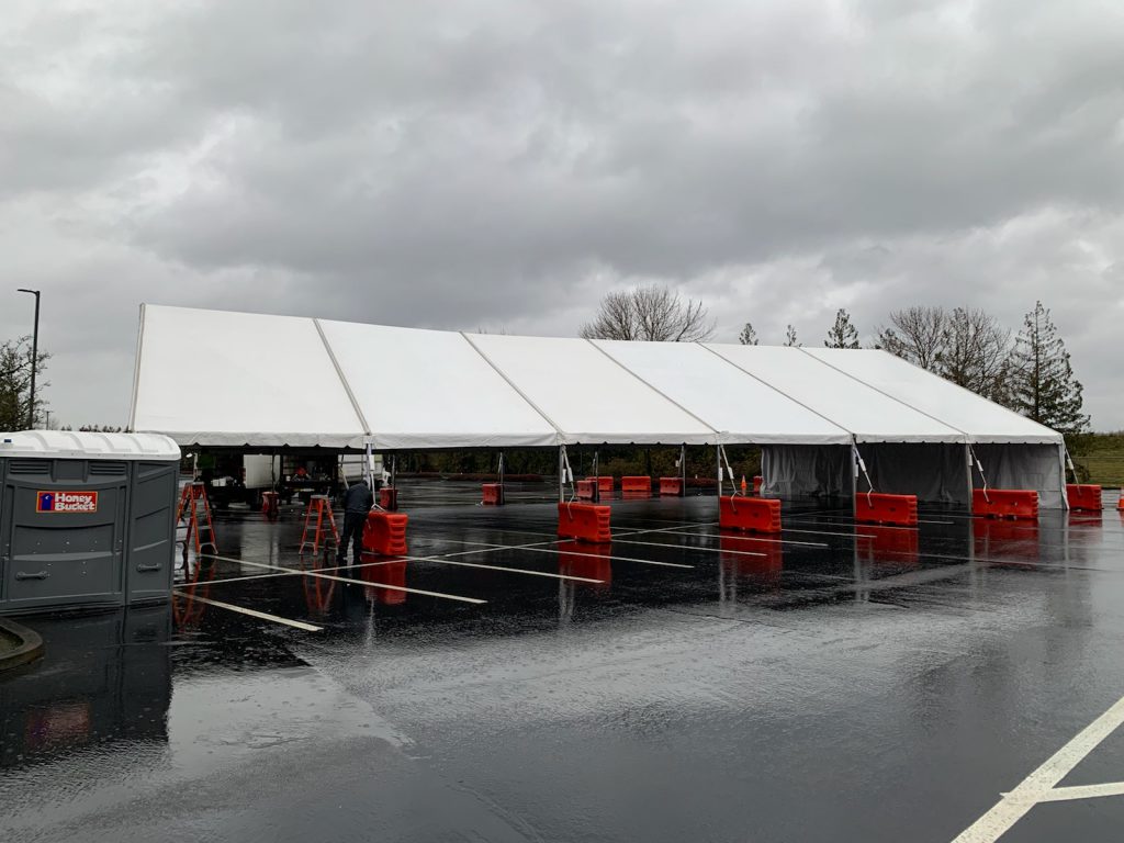 A photo of a big white tent with six rows, with rain puddles on the ground and dark clouds overhead.