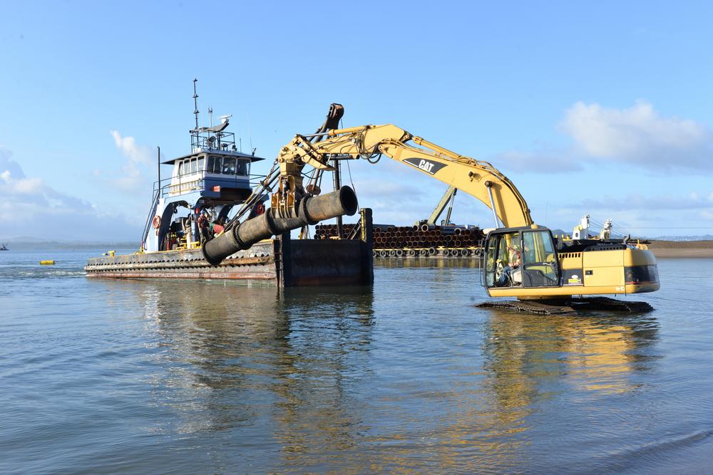 A crane sits in the water lifting a pipe onto a dredge vessel