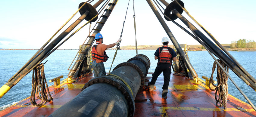 Two men stand next to a large pipe that sits on top of a ship. The pipe is pointing out toward the water